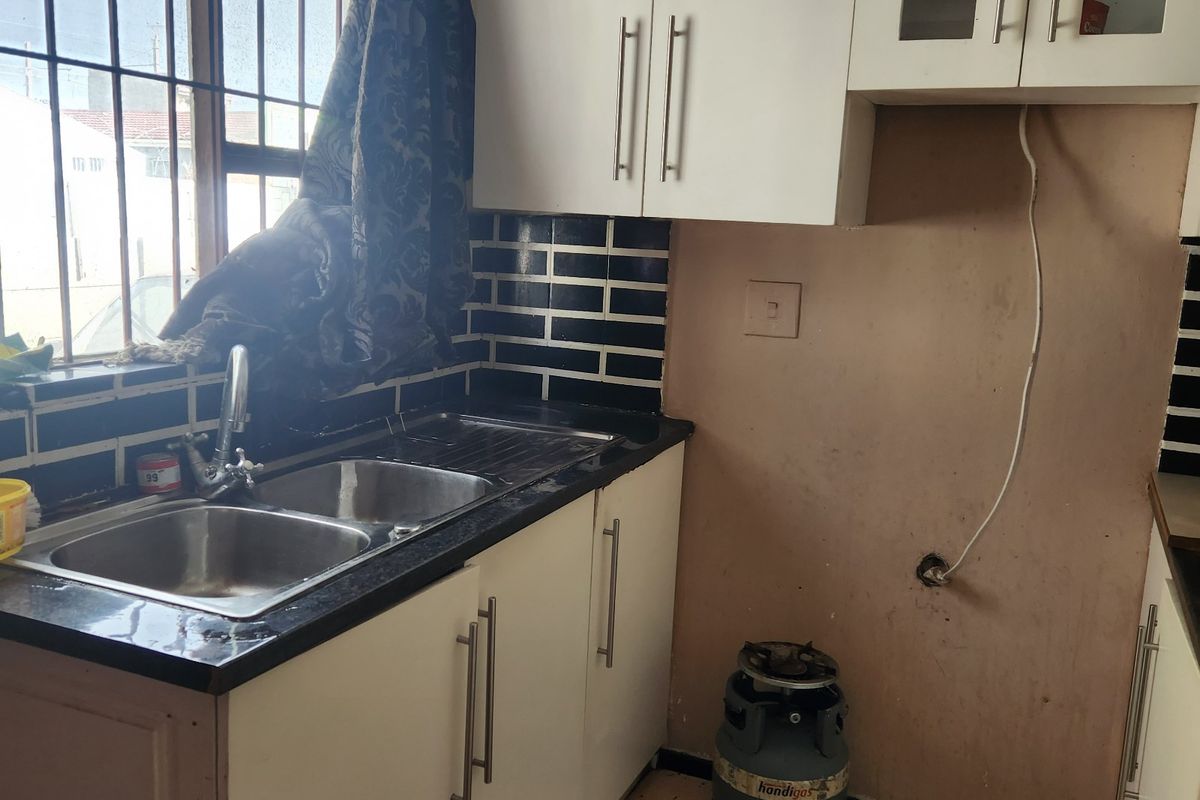 A compact kitchen featuring white cabinetry, a double bowl sink, and tiled splashbacks, currently requiring significant cosmetic updates and flooring repairs.