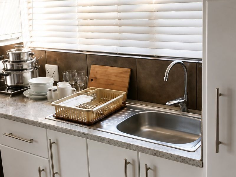 A functional kitchen area featuring white cabinetry, a stainless steel sink, tiled backsplash, and venetian blinds over the window.