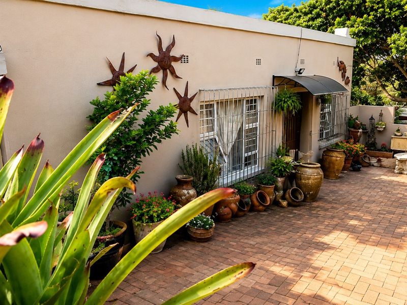 A paved outdoor patio area featuring a covered entrance, security bars on windows, and established garden surroundings.