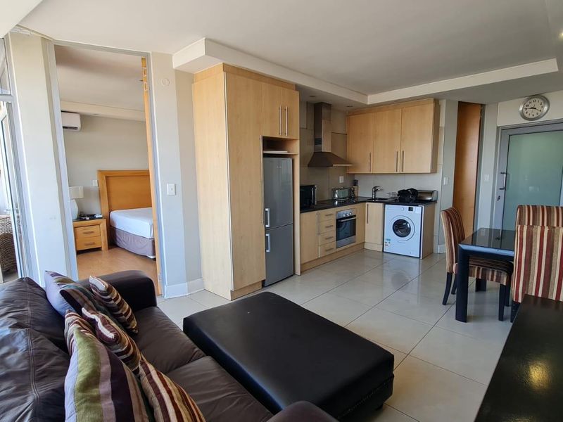 Open-plan living area featuring a compact kitchen with light wood cabinetry, integrated appliances, and tiled flooring, seamlessly connecting to the adjacent bedroom and balcony.