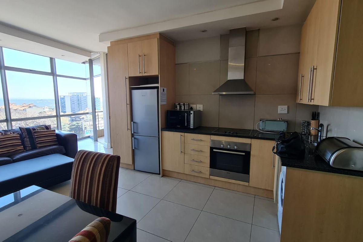Open-plan kitchen and living area featuring light wood cabinetry, integrated appliances, tiled flooring, and expansive windows offering natural light and urban views.