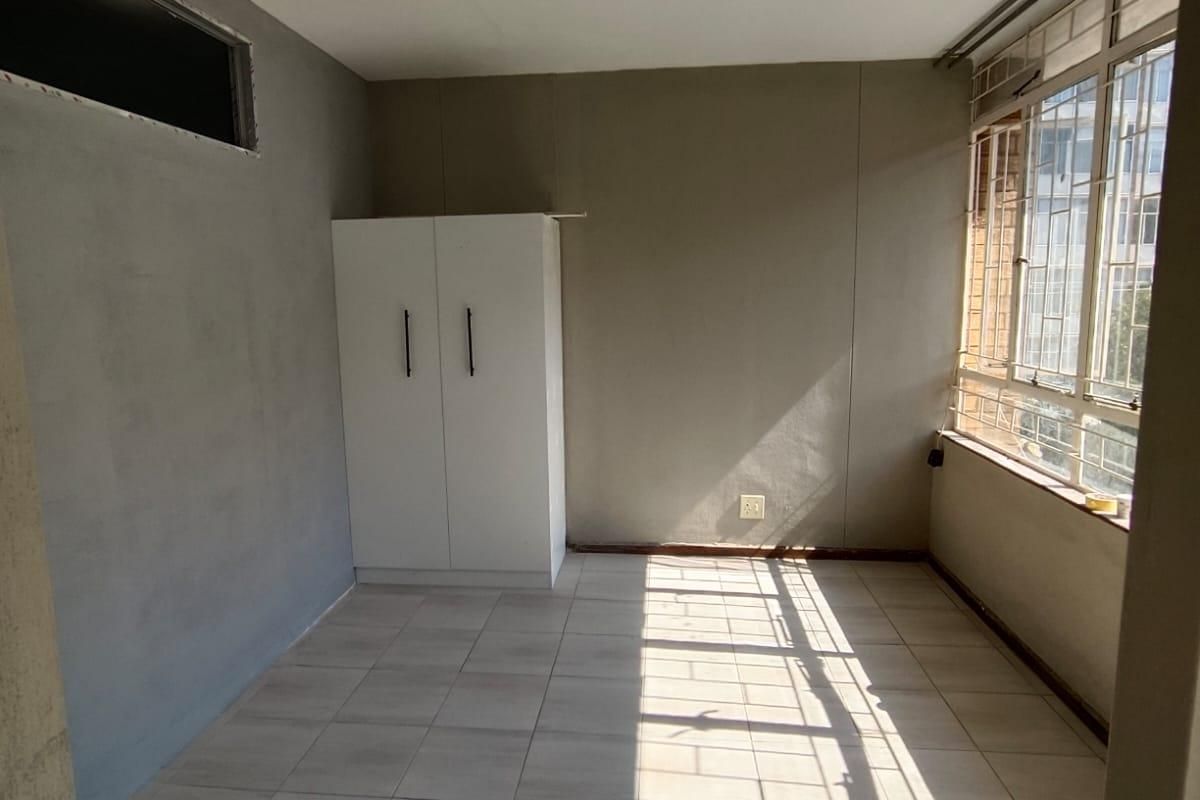 A vacant bedroom featuring tiled flooring, a built-in wardrobe, and large windows with security bars, currently requiring finishing touches to the wall plaster.