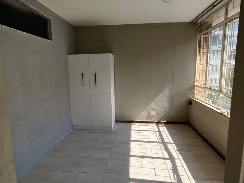 A vacant bedroom featuring tiled flooring, a built-in wardrobe, and large windows with security bars, currently requiring finishing touches to the wall plaster.