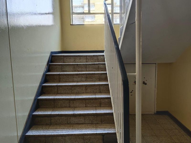 A functional apartment building stairwell featuring tiled steps, a metal balustrade, and a large window providing natural light to the common area.
