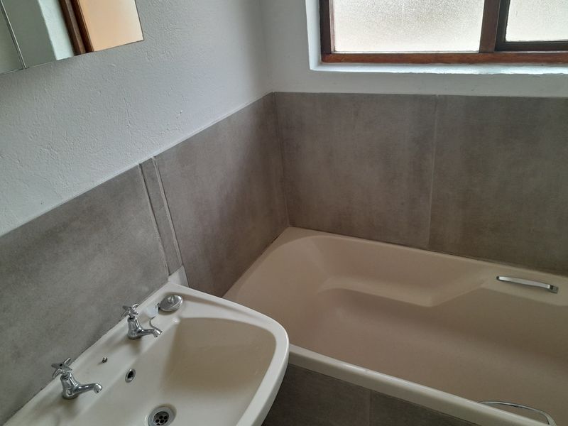 A functional bathroom featuring a neutral-toned bathtub and pedestal basin with modern grey tiled splashbacks and a timber-framed window.