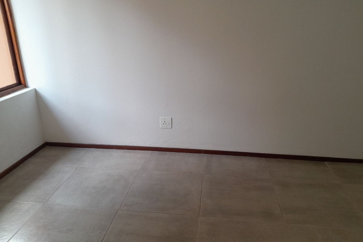 An empty bedroom featuring neutral tiled flooring, dark timber skirting boards, and a single electrical outlet on a plain wall.