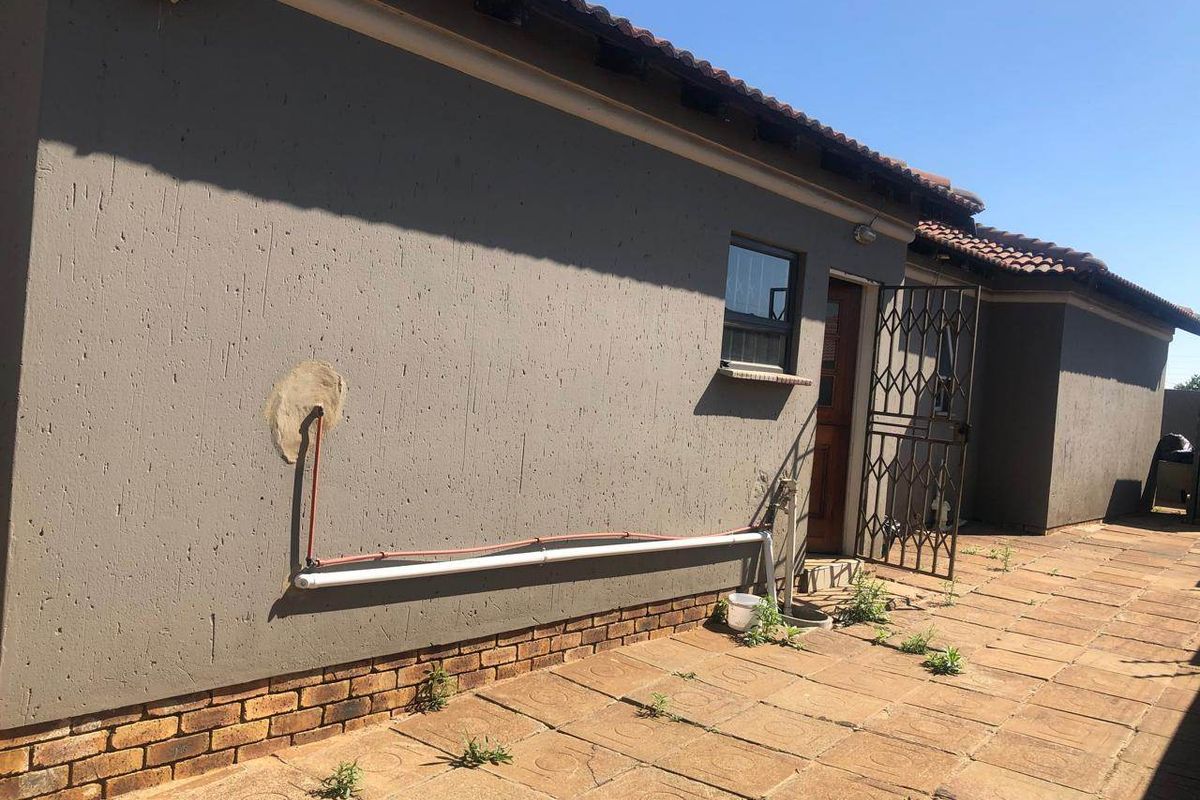 Rear exterior view of the property featuring a paved patio area, security gate, and exposed exterior plumbing along the wall.