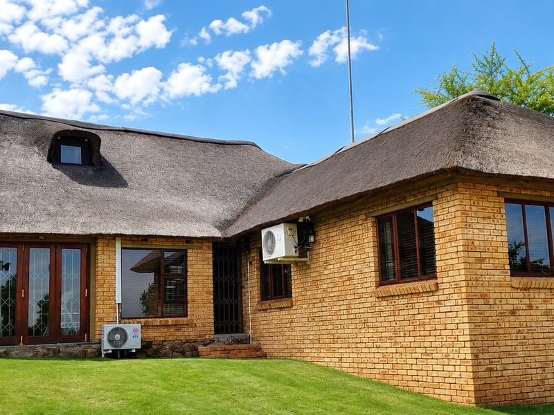 Rear view of a well-maintained face brick farmhouse featuring a traditional thatch roof, timber-framed windows, and folding doors opening onto a manicured lawn.