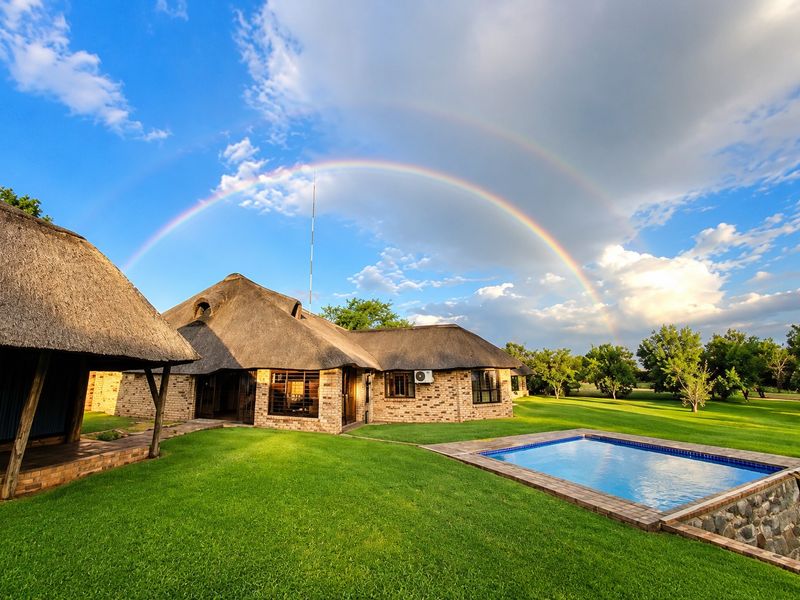 A scenic rear view of a farmhouse featuring a traditional thatch roof, brick exterior, and a private swimming pool set within a lush garden.