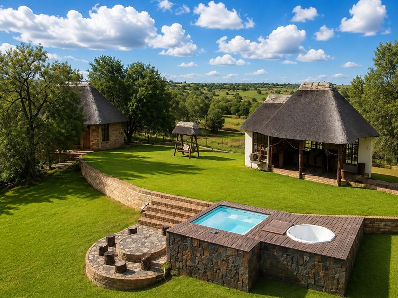 Aerial view of a farm property featuring a swimming pool, hot tub, timber decking, stone fire pit, and two thatched-roof structures set in landscaped grounds.