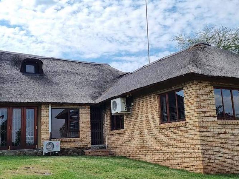 A well-maintained face brick farmhouse featuring a traditional thatch roof, timber-framed windows, and folding doors opening onto a landscaped garden area.