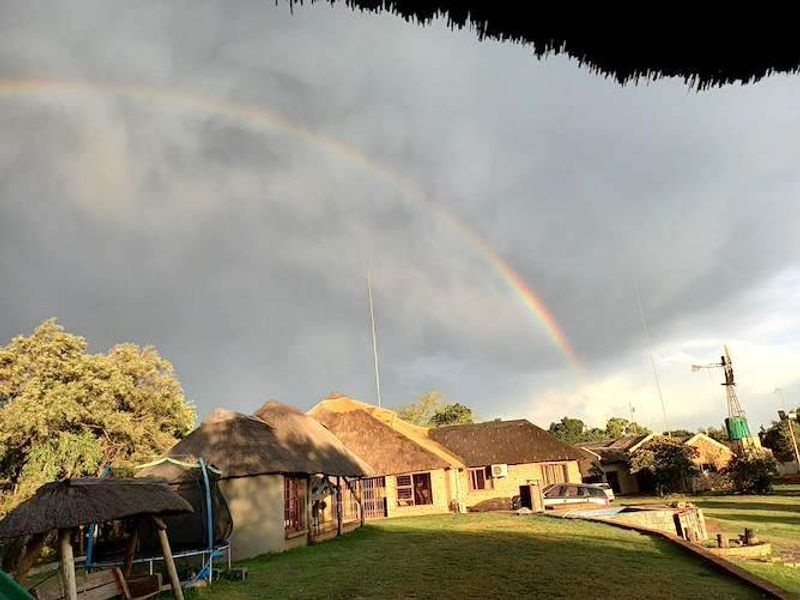 A rear view of a farmhouse featuring a traditional thatched roof, a swimming pool, and a windmill set against a scenic rainbow.
