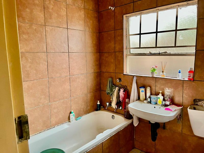 A dated bathroom featuring brown wall tiles, a standard bathtub, and a wall-mounted basin with a window providing natural light.
