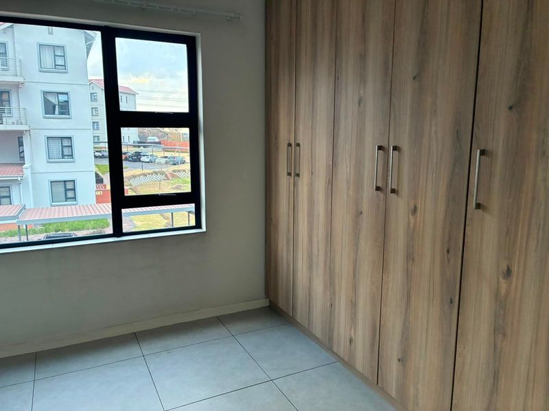A bright, empty bedroom featuring modern wood-finish built-in cupboards, large aluminium-framed windows, and neutral tiled flooring.