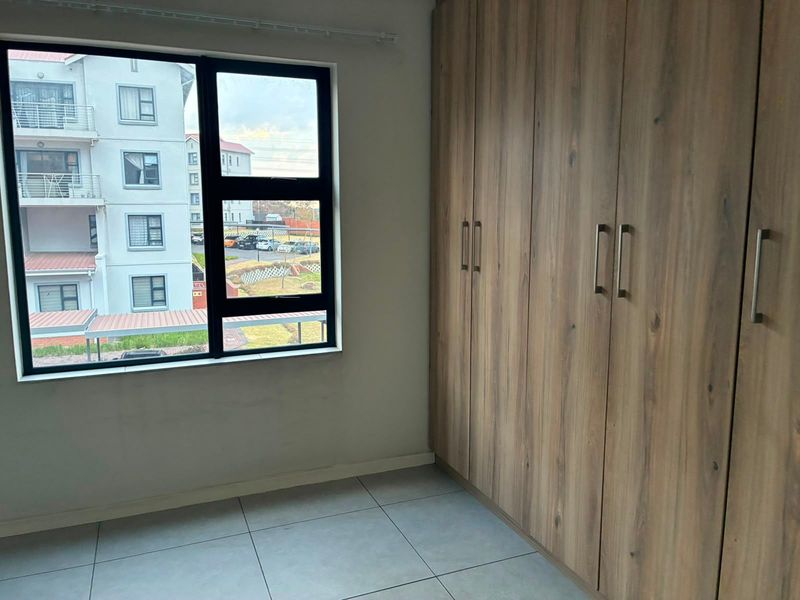 A bright, empty bedroom featuring modern wood-finish built-in cupboards, neutral tiled flooring, and a large window with views of the surrounding complex.