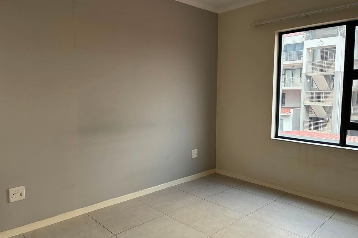 An empty bedroom featuring neutral tiled flooring, grey accent wall, and an aluminium-framed window with views of the adjacent building.