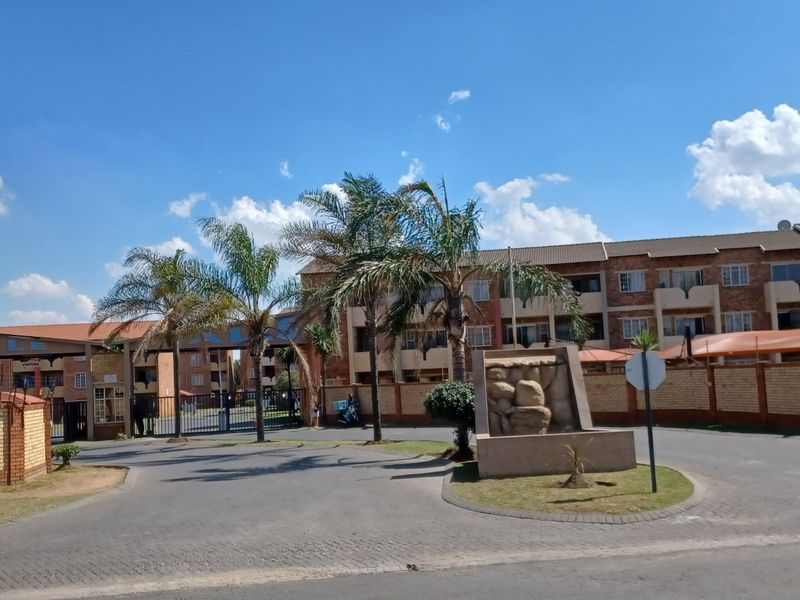 Front entrance view of the apartment complex featuring brick facade buildings, paved driveway, security gate, and perimeter wall with electric fencing.