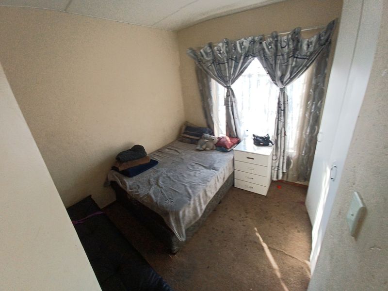 A compact bedroom featuring a built-in wardrobe and window with curtains. The room requires cosmetic updates to the flooring and wall finishes.