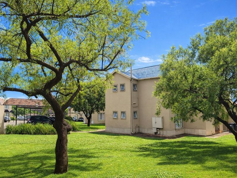 A view of the apartment complex exterior featuring a well-maintained lawn, mature trees, and a multi-story building with a tiled roof.