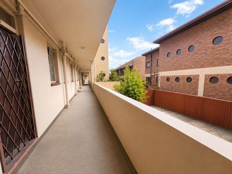 An open-air apartment balcony walkway featuring a solid balustrade, concrete flooring, and access to residential units with security gates.