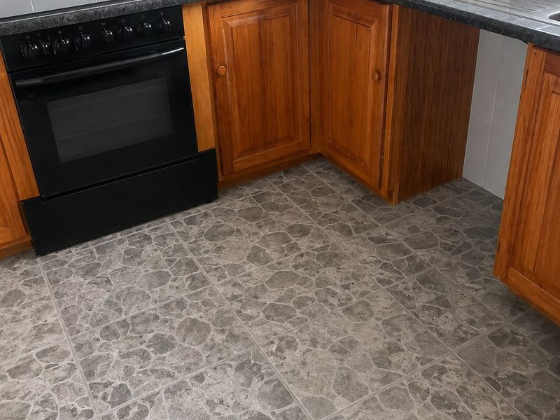A functional kitchen featuring wooden cabinetry, a built-in oven with hob, granite-look countertops, and a stainless steel sink with tiled splashback.
