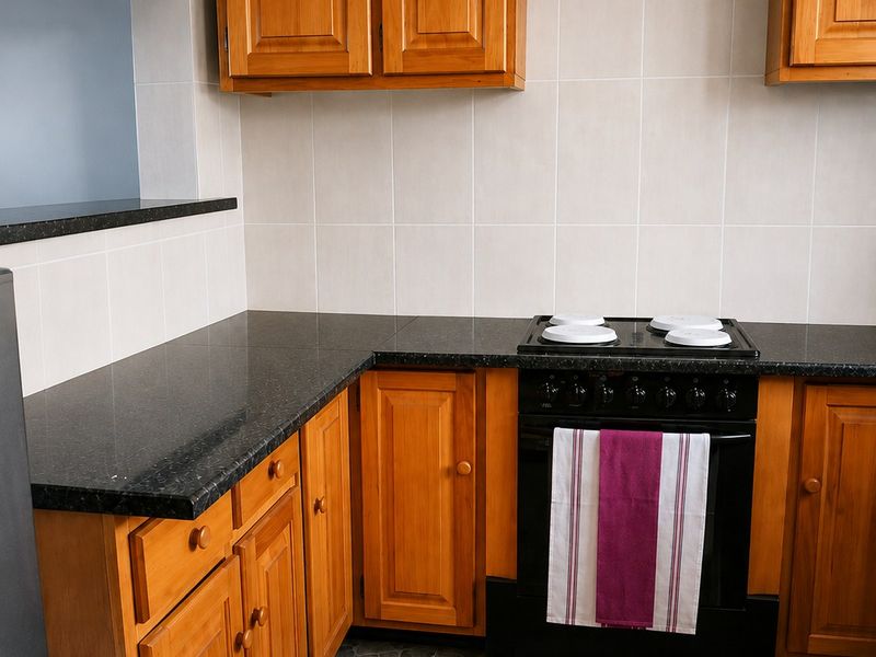 A functional kitchen featuring warm wooden cabinetry, dark granite countertops, a built-in oven with electric hob, and durable stone-look floor tiles.