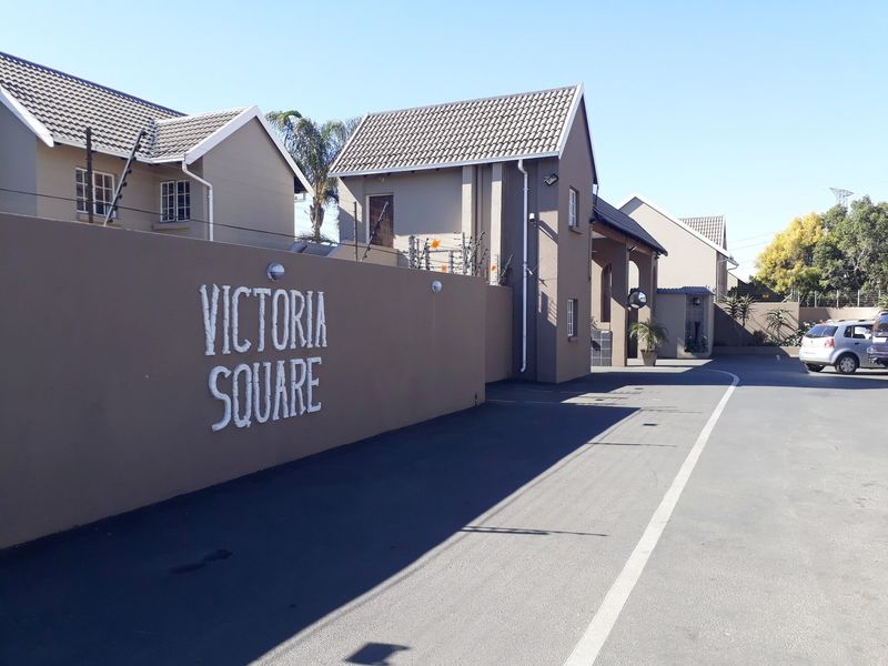 Exterior view of the Victoria Square townhouse complex entrance, featuring a paved driveway, perimeter wall with electric fencing, and tiled roof structures.