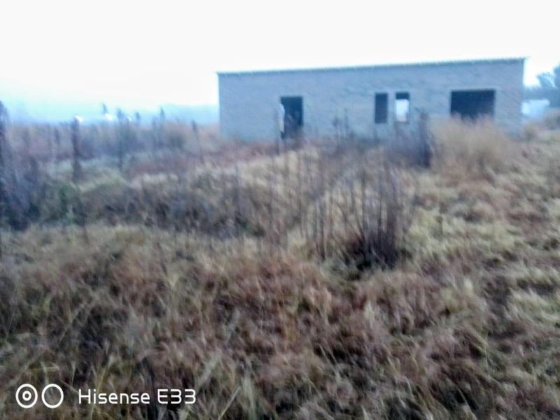 A view of an unfinished masonry structure situated on a large, overgrown vacant residential plot.
