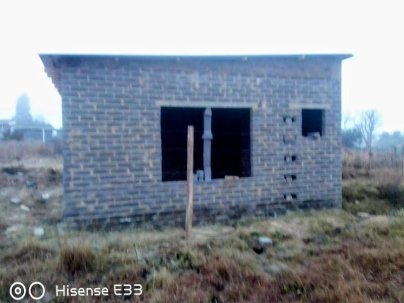 An unfinished residential structure featuring exposed brickwork and open window frames, situated on a vacant plot of land.
