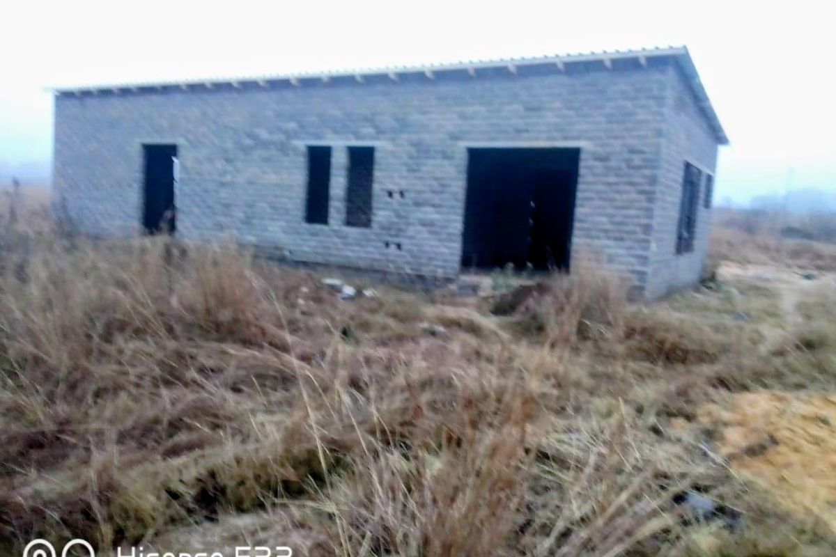 An unfinished residential structure featuring concrete block walls and a metal roof, situated on a vacant plot with overgrown grass.