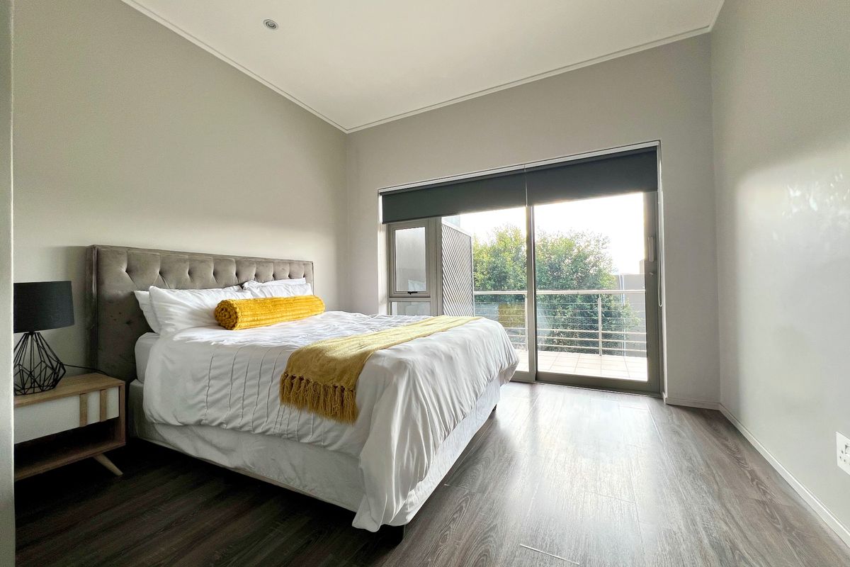 A bright bedroom featuring modern laminate flooring, neutral walls, and a large sliding glass door providing access to a private balcony.