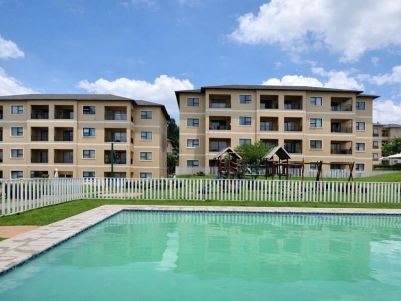 A well-maintained communal swimming pool area featuring a picket fence, landscaped lawn, and playground equipment set against apartment building facades.