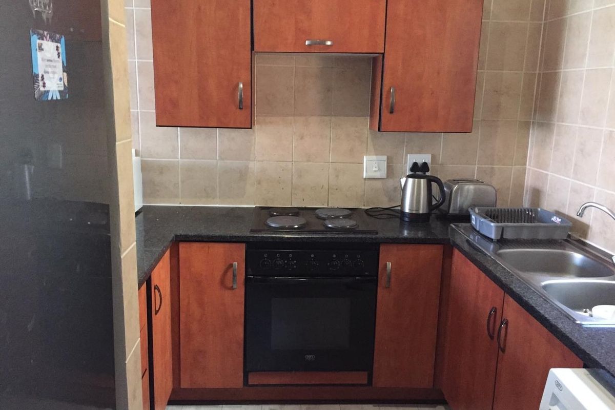A compact kitchen featuring wooden cabinetry, dark laminate countertops, a built-in oven with electric hob, and a double bowl sink with tiled backsplash.