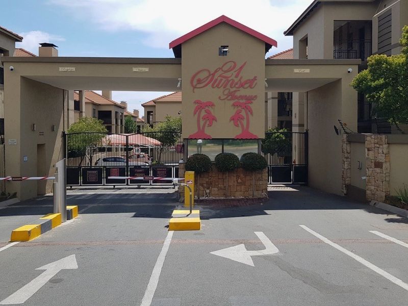 Secure gated entrance to the apartment complex featuring a boom gate, stone-clad pillars, and clear signage for Sunset Avenue.
