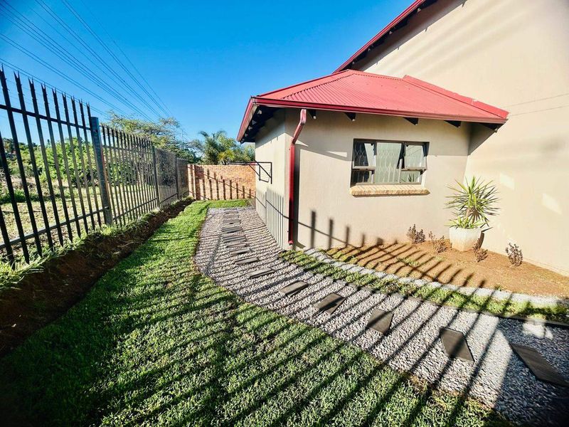 A well-maintained rear exterior area featuring a stone pathway, manicured lawn, metal security fencing, and a clean building facade with a red metal roof.