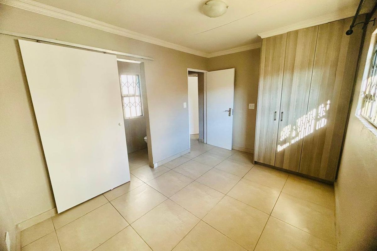 A clean, empty bedroom featuring neutral tiled flooring, built-in wooden cupboards, and security bars on the windows.