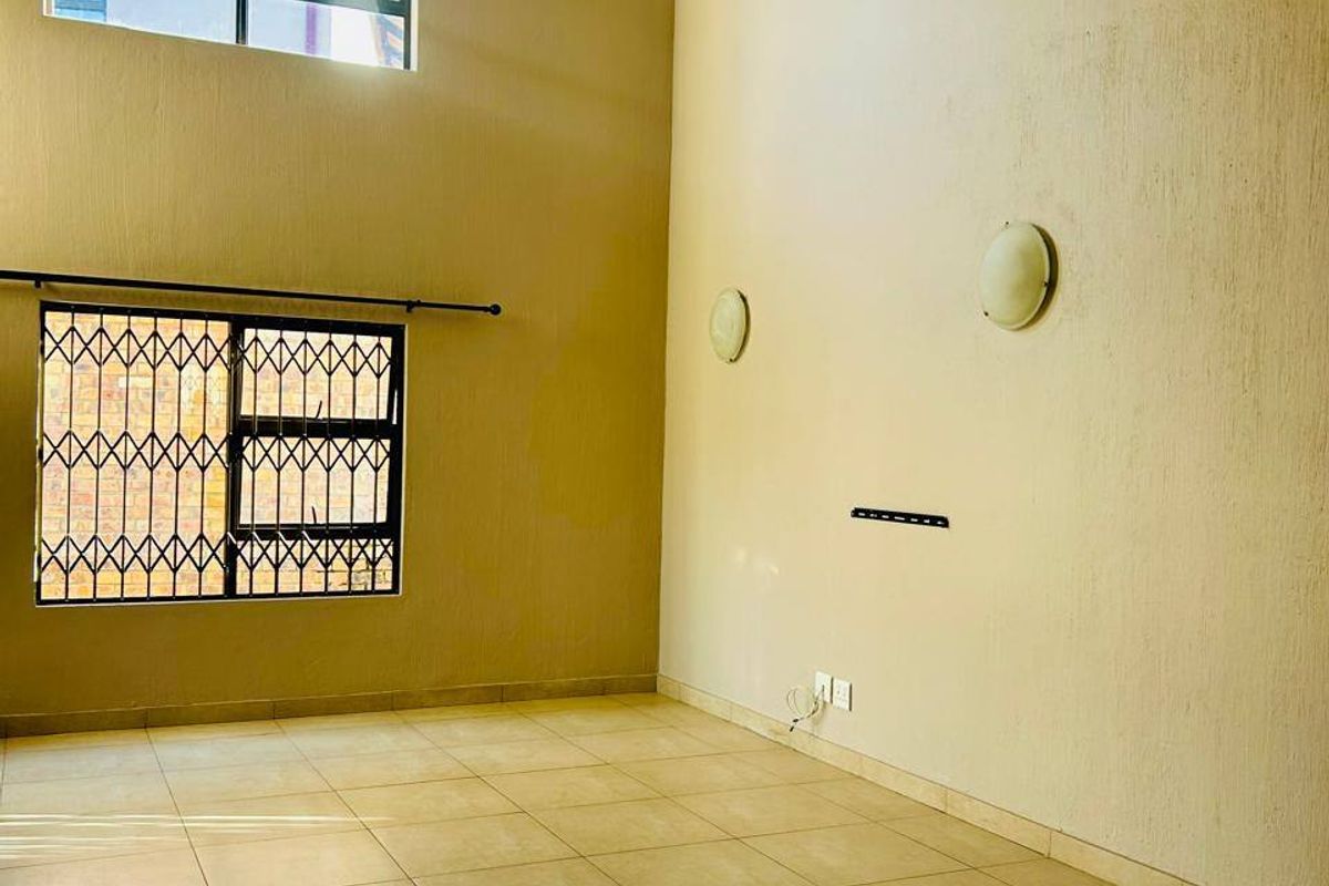 Spacious double-volume living area featuring tiled flooring, large windows with security bars, and wall-mounted lighting, offering a bright and open blank canvas.