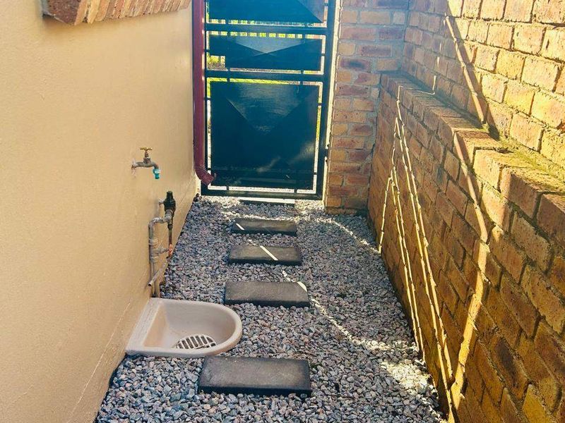 A side passage of the townhouse featuring a gravel pathway with stepping stones, a brick boundary wall, and a secure metal gate.