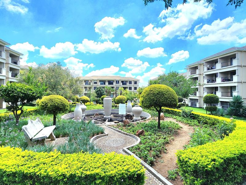 A well-maintained communal garden area featuring a central water feature, manicured hedges, and paved pathways surrounded by apartment building facades.