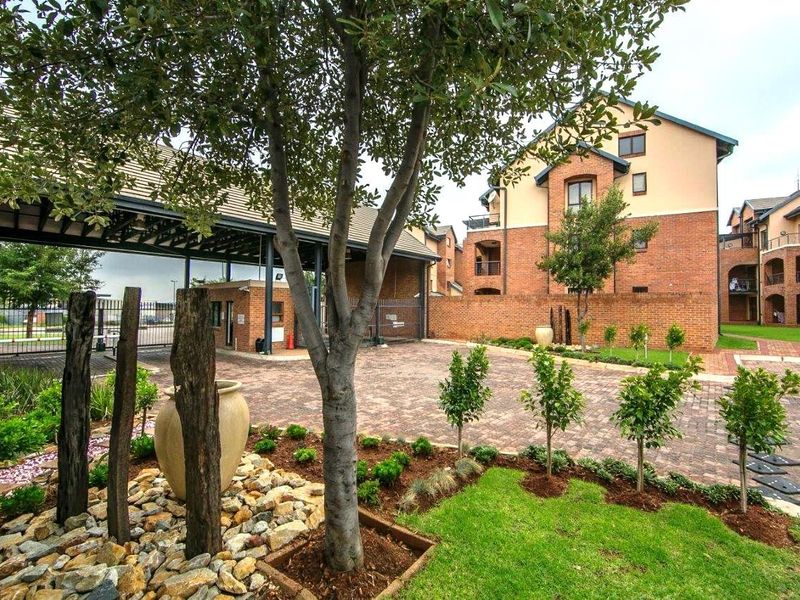 View of the apartment complex entrance featuring a secure gated access point, brick paved driveway, and well-maintained landscaped garden areas.