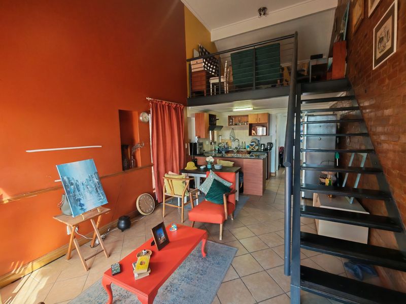 Open-plan living area featuring a mezzanine level, exposed brick walls, tiled flooring, and a functional kitchen space with a metal staircase.