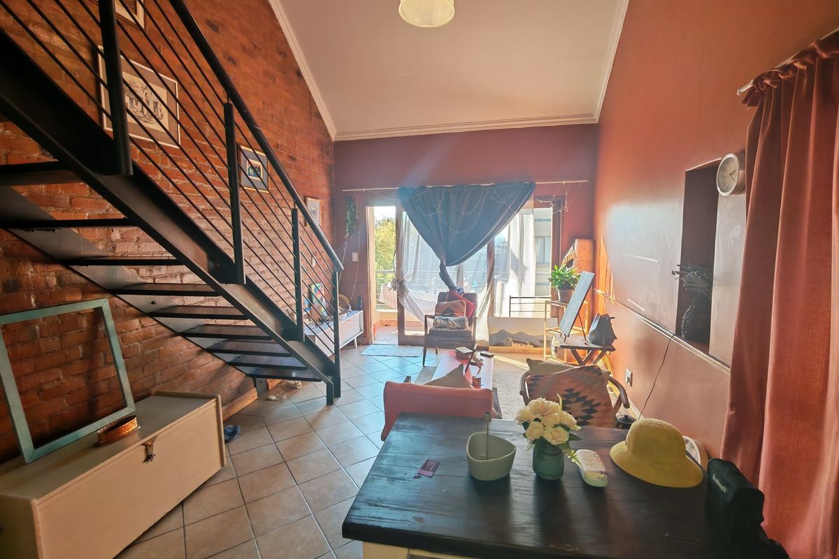 Spacious living area featuring an exposed brick wall, vaulted ceiling, and a metal staircase leading to an upper level, with access to a balcony.