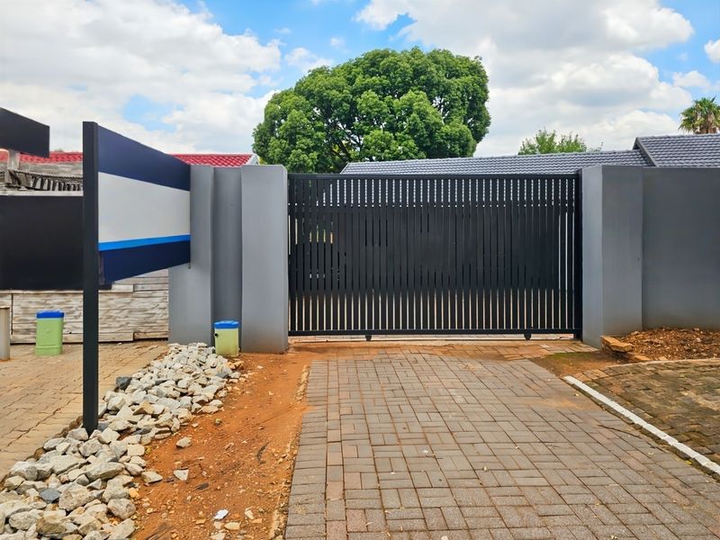 Front entrance featuring a modern black sliding gate, concrete boundary walls, and a paved driveway with adjacent unfinished landscaping.