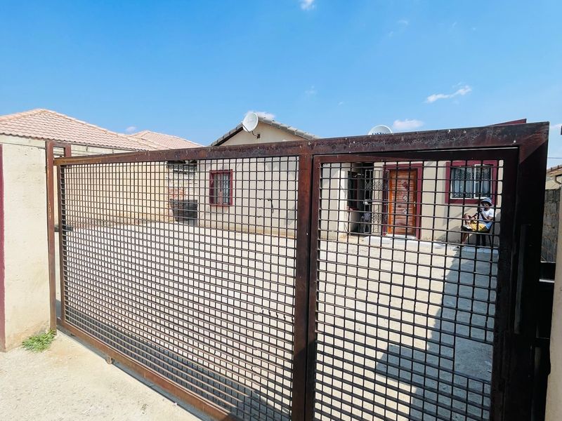 Front view of the property featuring a large metal security gate, concrete driveway, and a single-story house with tiled roofing and window security bars.