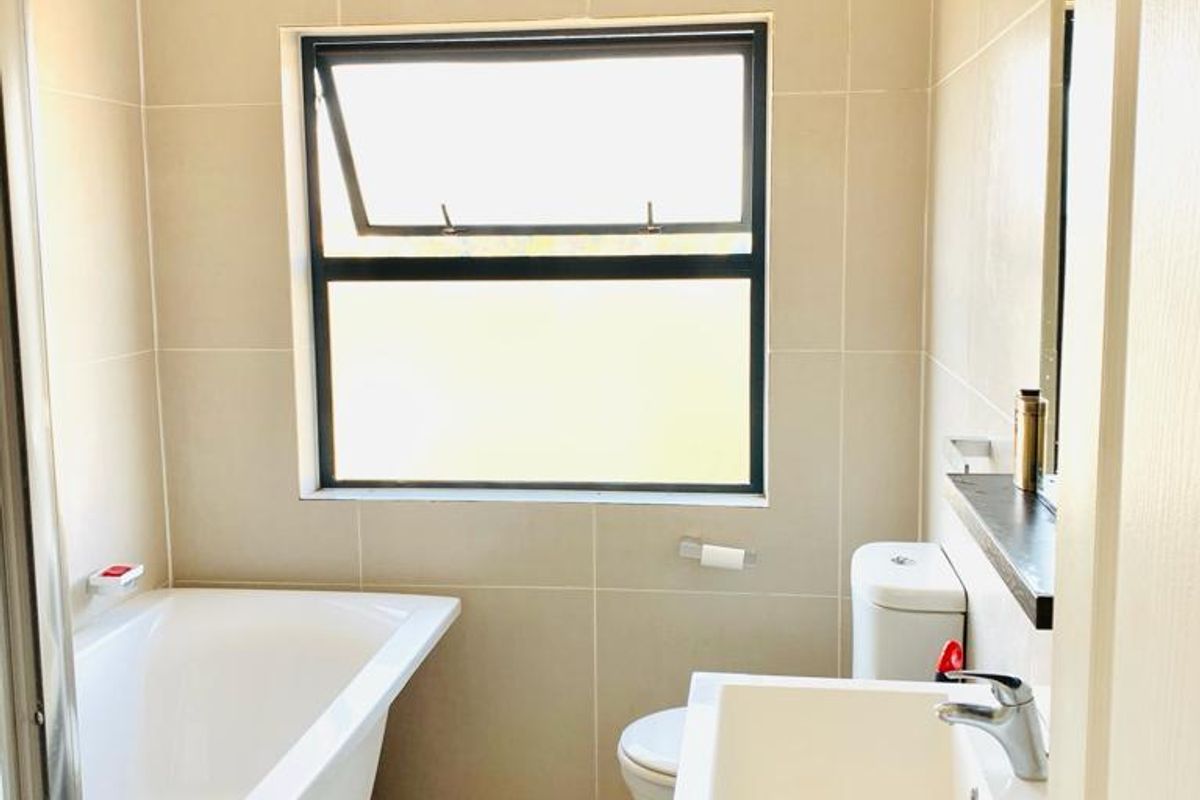 A clean, modern bathroom featuring neutral wall tiles, a white bathtub, a vanity basin, and a toilet, with natural light from a top-hung window.