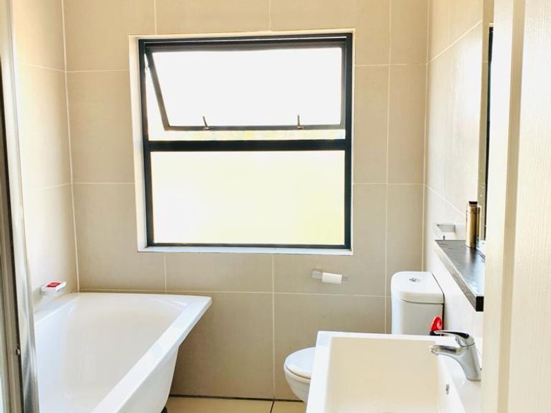 A clean, modern bathroom featuring neutral wall tiles, a white bathtub, a vanity basin, and a toilet, with natural light from a top-hung window.