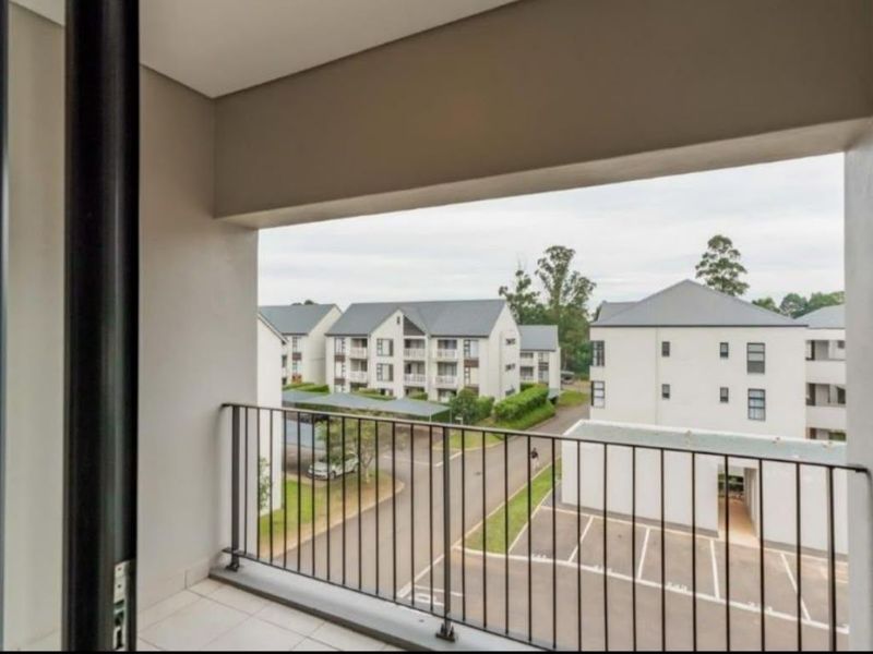 A clean, covered balcony featuring a metal balustrade and tiled flooring, offering an elevated view of the surrounding apartment complex and parking area.