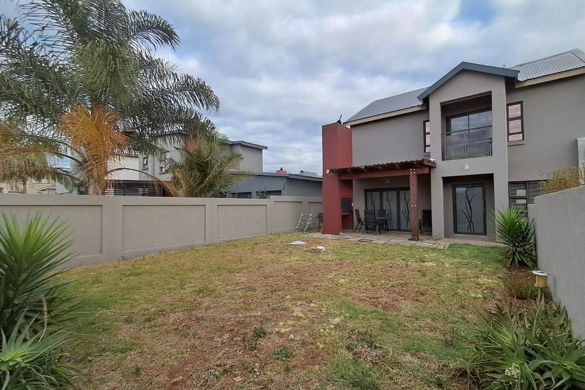 Rear view of a two-story house featuring a covered patio with a built-in braai, a small balcony, and a private garden with perimeter walling.
