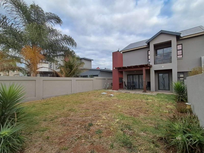 Rear view of a two-story house featuring a covered patio with a built-in braai, a small balcony, and a private garden with perimeter walling.