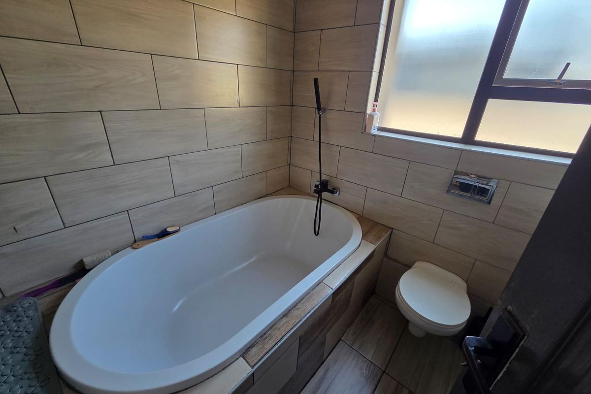 A modern bathroom featuring a built-in bathtub with wall-mounted shower fixture, a toilet, and neutral wood-look wall and floor tiling.